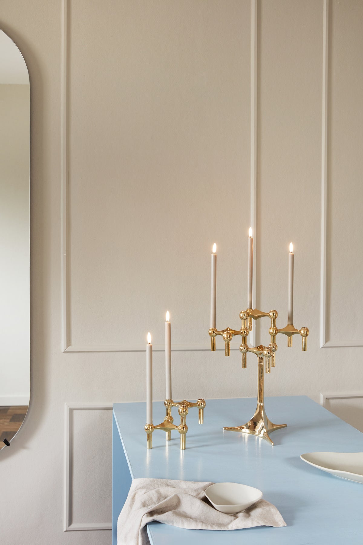 STOFF Nagel brass stand and a lower holder grouping with lit taper candles on a blue table in a wall paneled dining room
