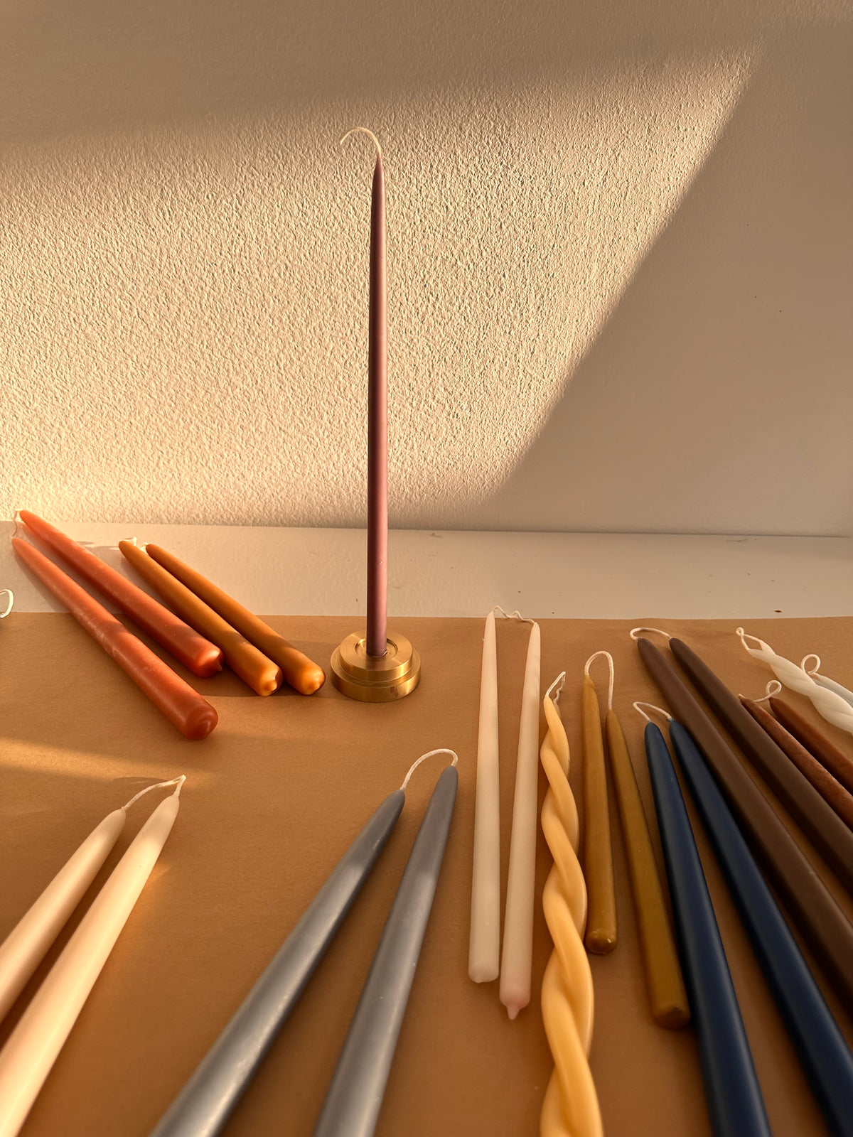 A slender pink GLŌAM Beeswax Taper standing in a solid brass candle holder, surrounded by hand-dipped candles in warm neutrals, ochre, and blue.