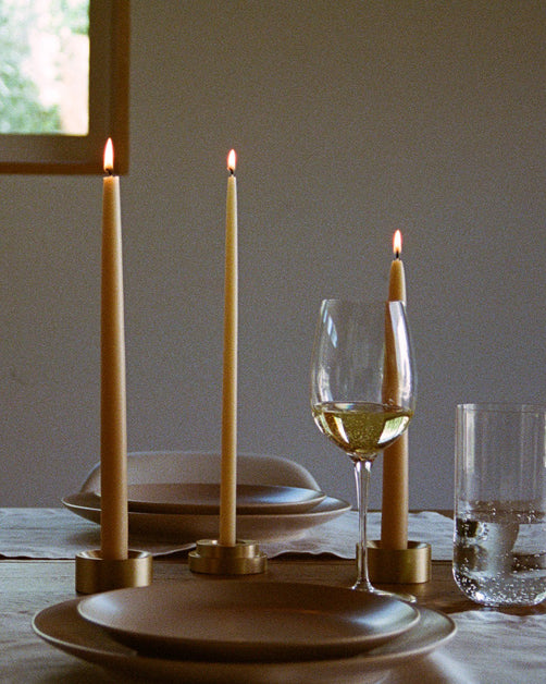 Lit GLŌAM Classic Beeswax Tapers in brass candle holders on a dining table set with ceramic plates, linen, and glassware.