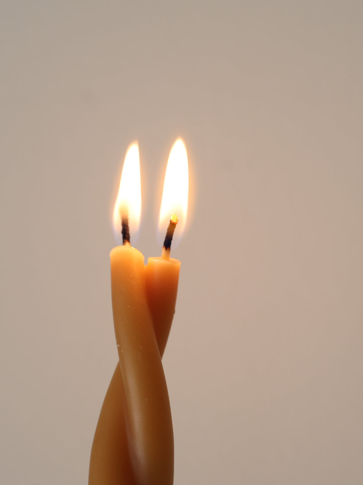 Close-up of a lit GLŌAM Twisted Beeswax Taper with two flames rising from its intertwined candle form against a warm neutral background.