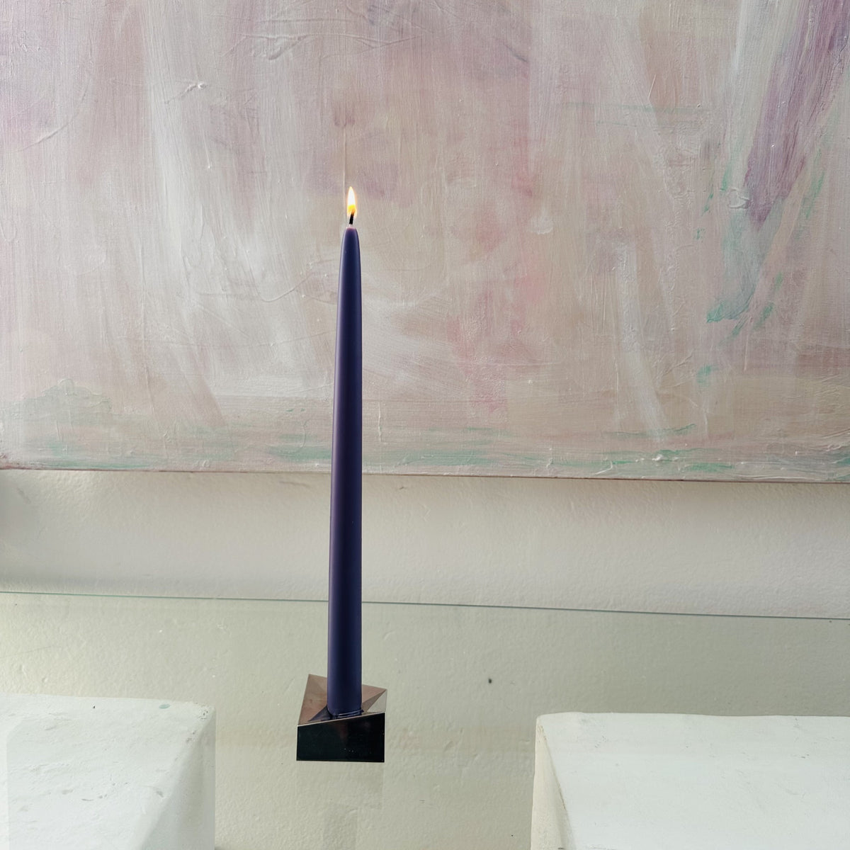 A lit Maritime Blue beeswax taper candle in a geometric brass holder, placed against a pale abstract painting.