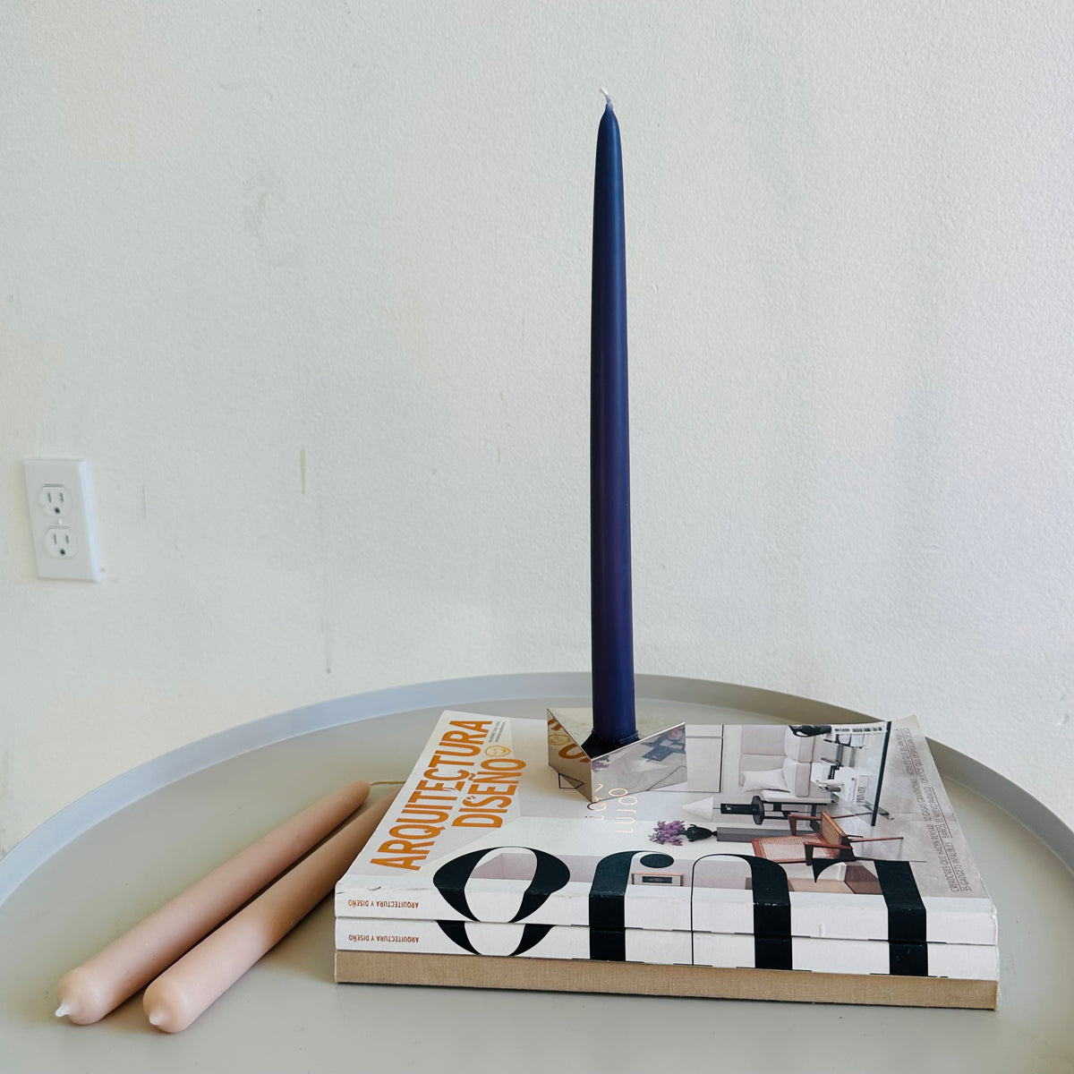 Maritime Blue beeswax taper candle in a brass holder on architecture magazines, with pale pink candles on a round table.
Copy alt text