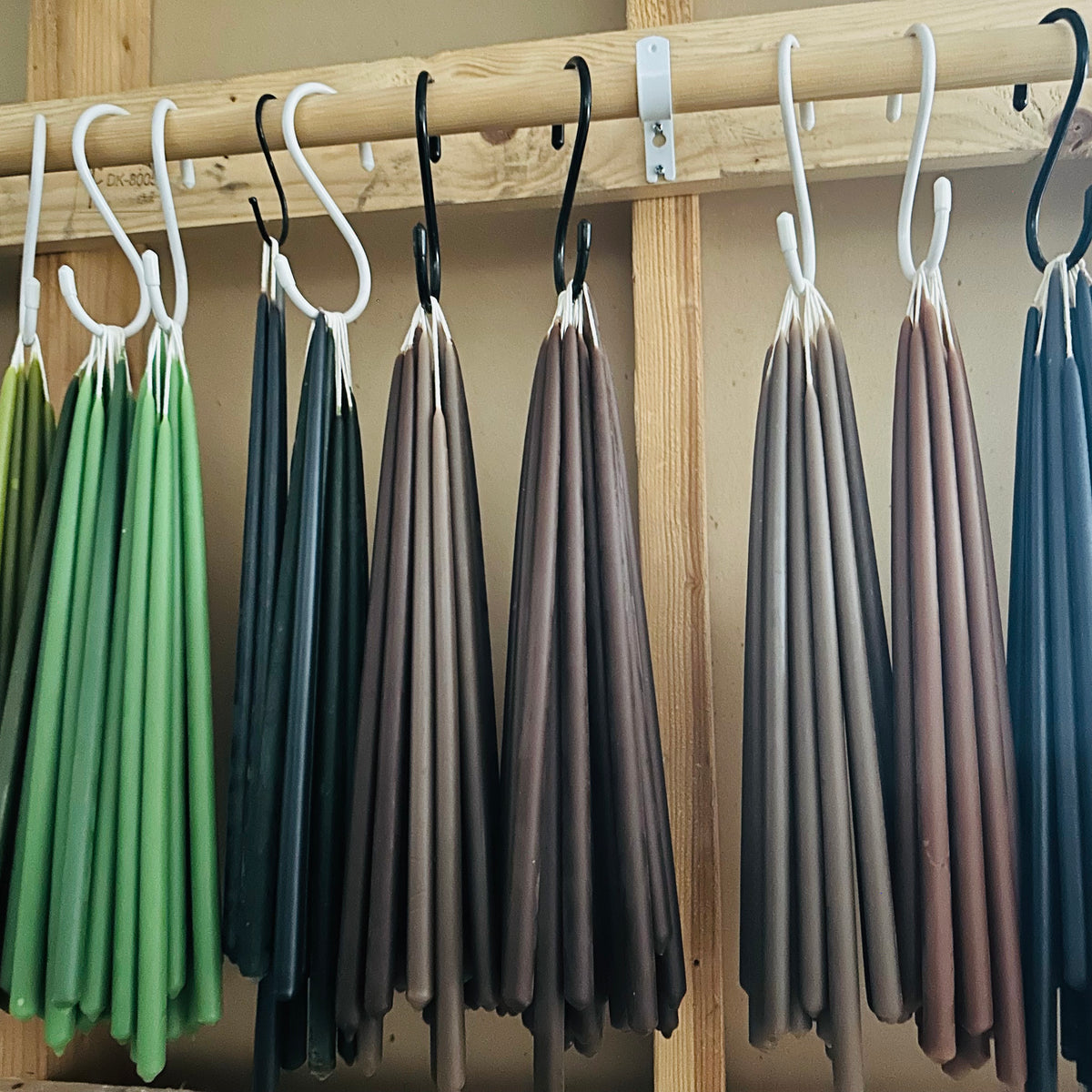 Hand-dipped GLŌAM Classic Beeswax Tapers in muted greens, mineral greys, browns, and blues hanging from a wooden rack in the studio.