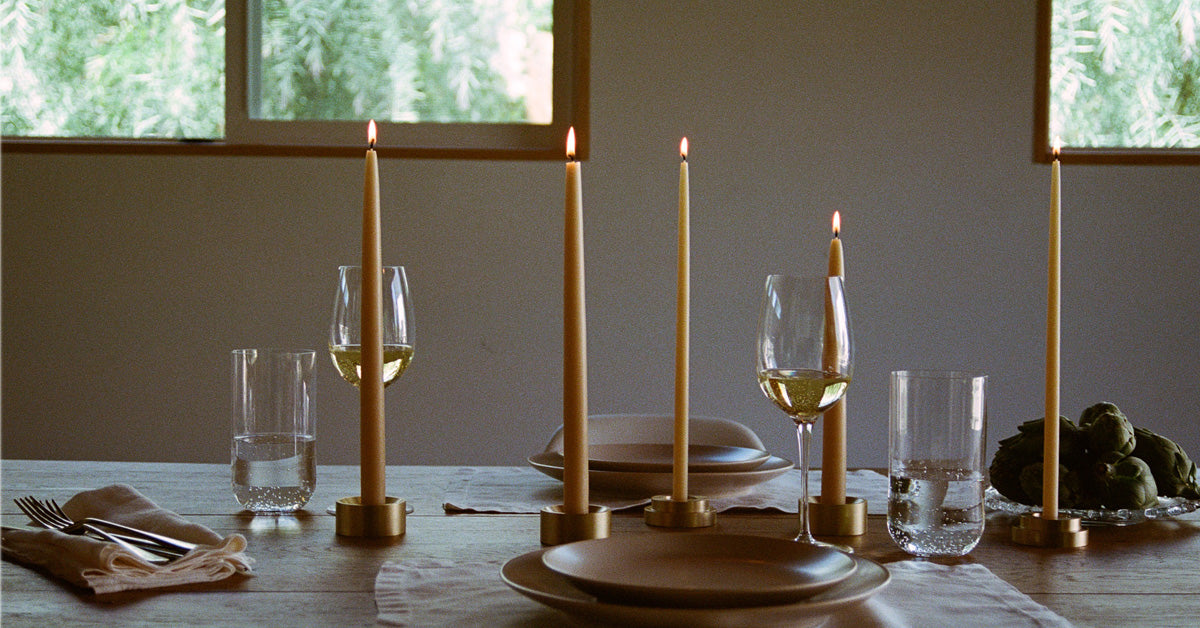 A styled dining table setting with tall taper candles in brass holders, wine glasses, plates, folded linen napkins with cutlery, and artichokes, photographed indoors near windows with soft natural light.