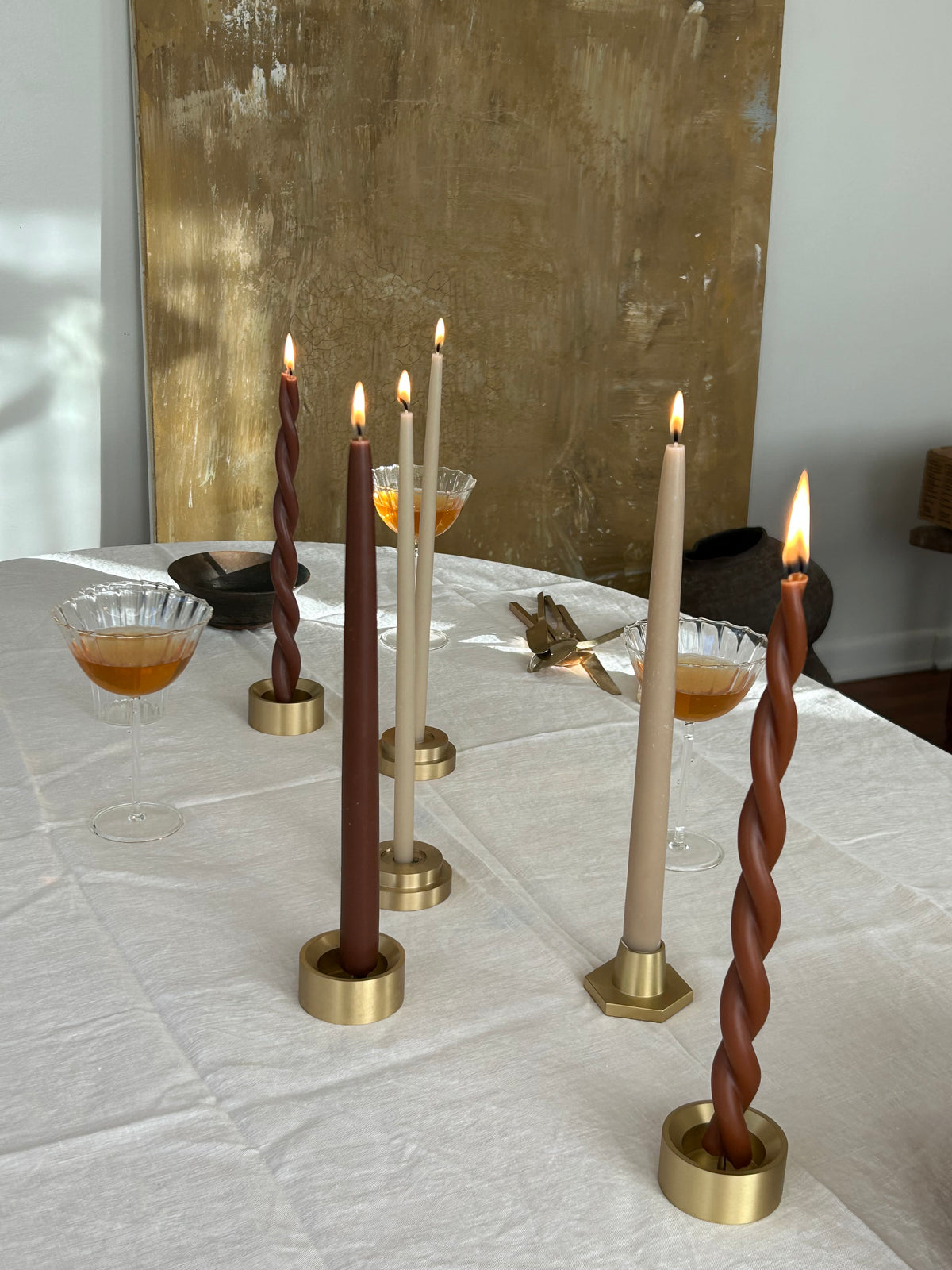 GLŌAM Classic and Twisted Beeswax Tapers burning in solid brass candle holders on a white linen-covered table with glassware and a textured backdrop.