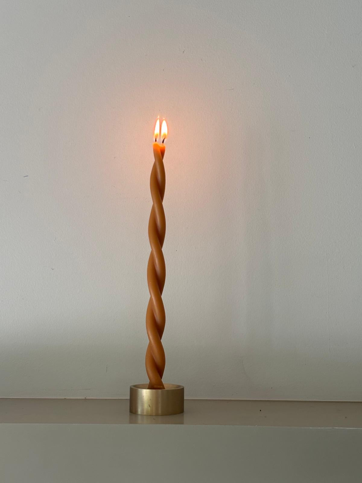 A single lit GLŌAM Twisted Beeswax Taper standing in a small brass candle holder against a pale grey wall.
