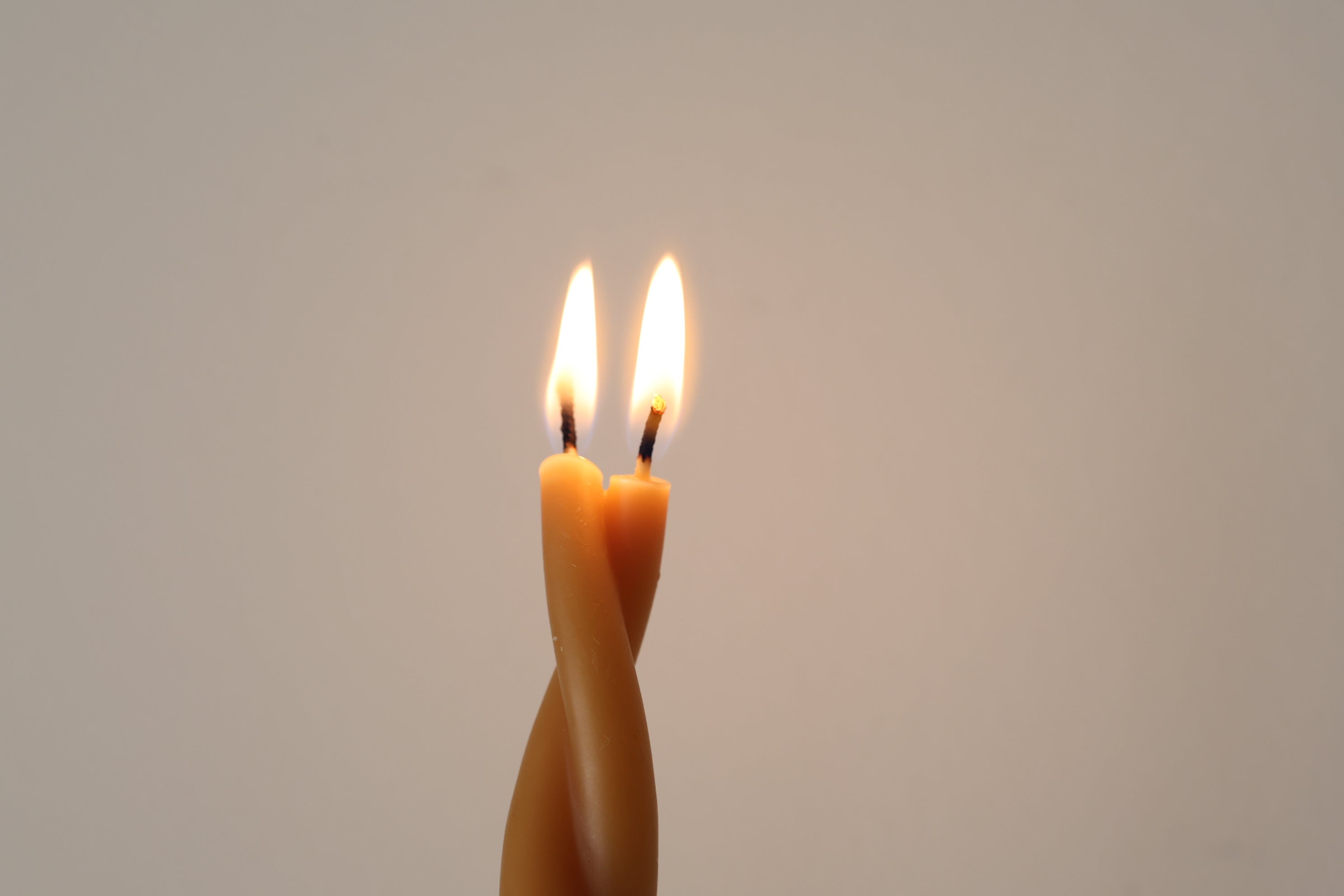 Close-up of a lit GLŌAM Twisted Beeswax Taper with two flames rising from its intertwined candle form against a warm neutral background.