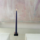 A lit Maritime Blue beeswax taper candle in a geometric brass holder, placed against a pale abstract painting.