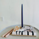 Maritime Blue beeswax taper candle in a brass holder on architecture magazines, with pale pink candles on a round table.
 Copy alt text