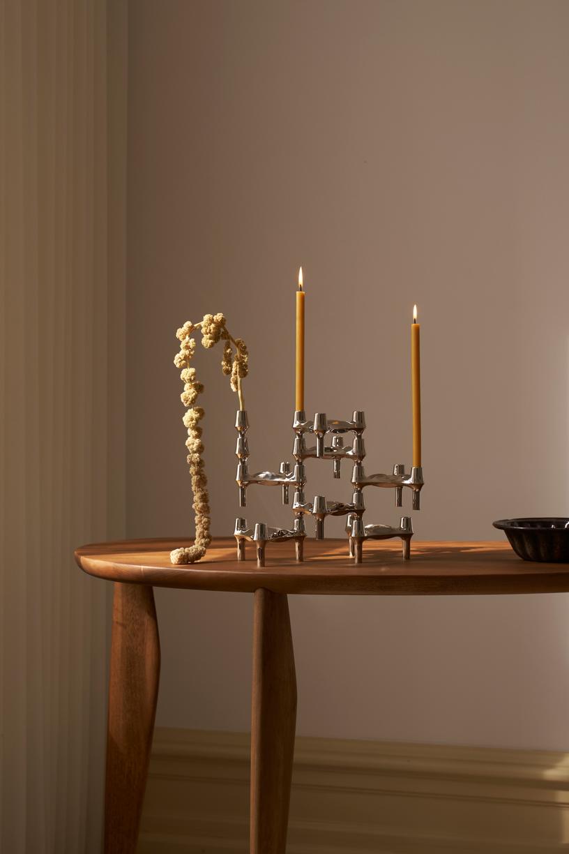 STOFF Nagel BMF chrome candle holders built into a composition on a wooden table, holding two lit taper candles beside a dried branch