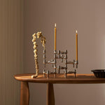STOFF Nagel BMF chrome candle holders built into a composition on a wooden table, holding two lit taper candles beside a dried branch