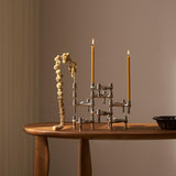 STOFF Nagel BMF chrome candle holders built into a composition on a wooden table, holding two lit taper candles beside a dried branch