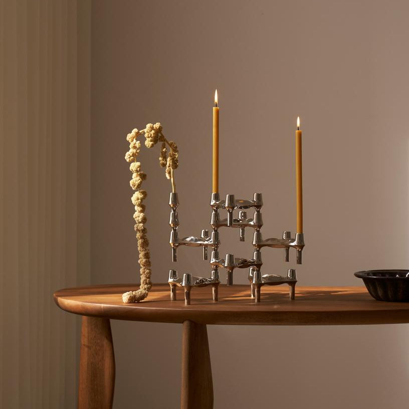 STOFF Nagel BMF chrome candle holders built into a composition on a wooden table, holding two lit taper candles beside a dried branch