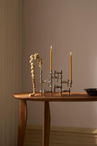 STOFF Nagel BMF chrome candle holders built into a composition on a wooden table, holding two lit taper candles beside a dried branch
