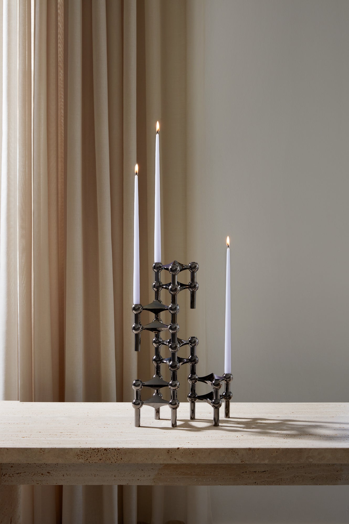 STOFF Nagel black candle holders holding three lit taper candles on a travertine surface against a soft curtain