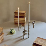 STOFF Nagel candle holders in chrome holding lit taper candles, styled with stacked books and a white flower in warm light