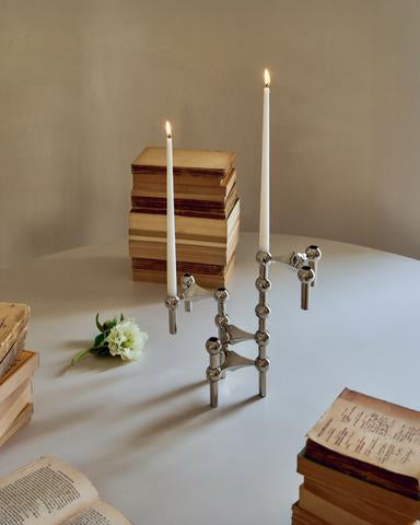 STOFF Nagel candle holders in chrome holding lit taper candles, styled with stacked books and a white flower in warm light