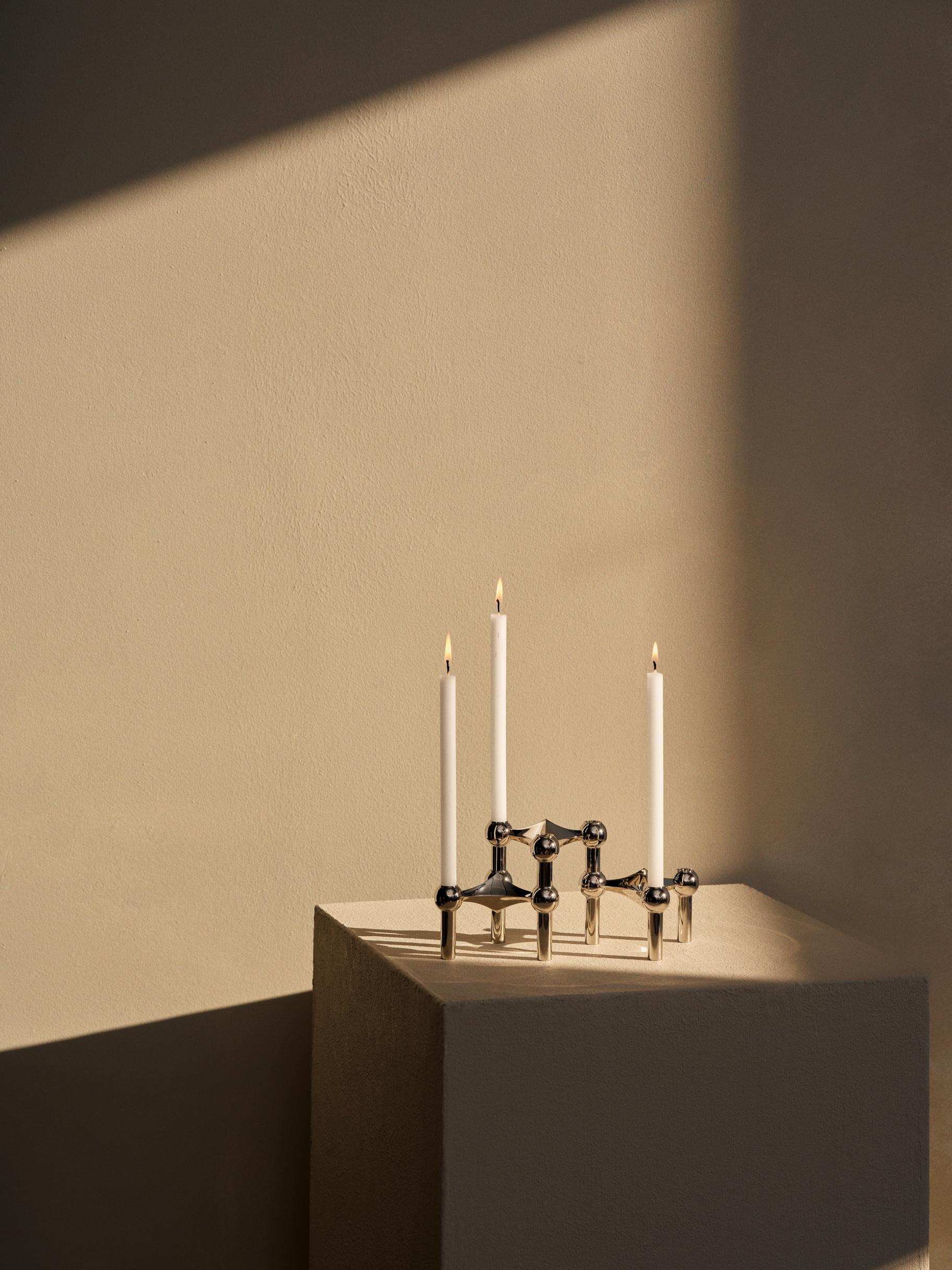 : STOFF Nagel chrome candle holders on a plinth holding three lit taper candles in a sunlit interior