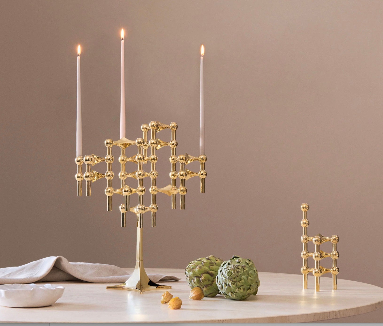 STOFF Nagel stand in solid brass built up with modular holders and lit taper candles, styled on a table with artichokes and linen